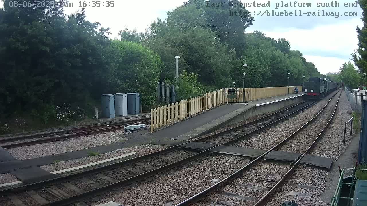 A dark-colored steam train is departing a train station on a partly cloudy day, surrounded by lush green trees.
