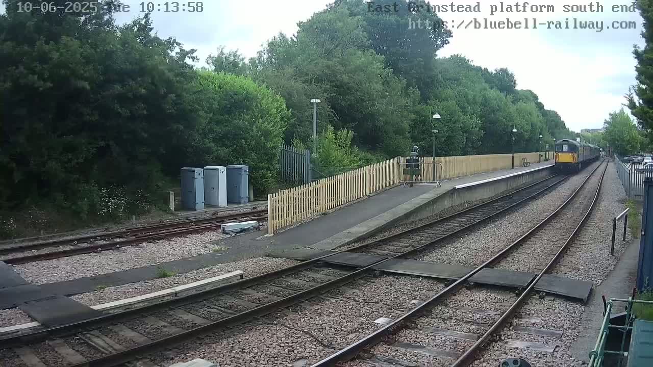 A train departs a railway station platform under partly cloudy skies, passing alongside a wooden fence and several gray utility boxes.