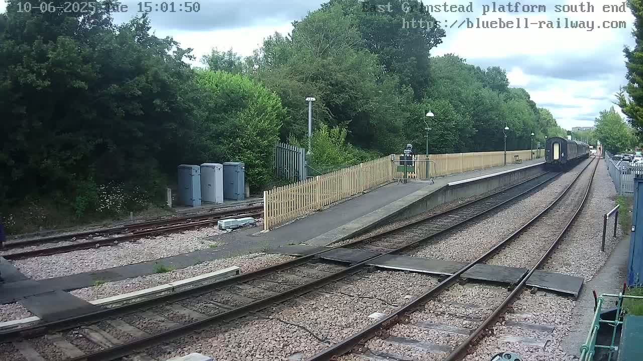 A train is departing from a railway platform on an overcast day, passing by a wooden fence and several utility boxes.