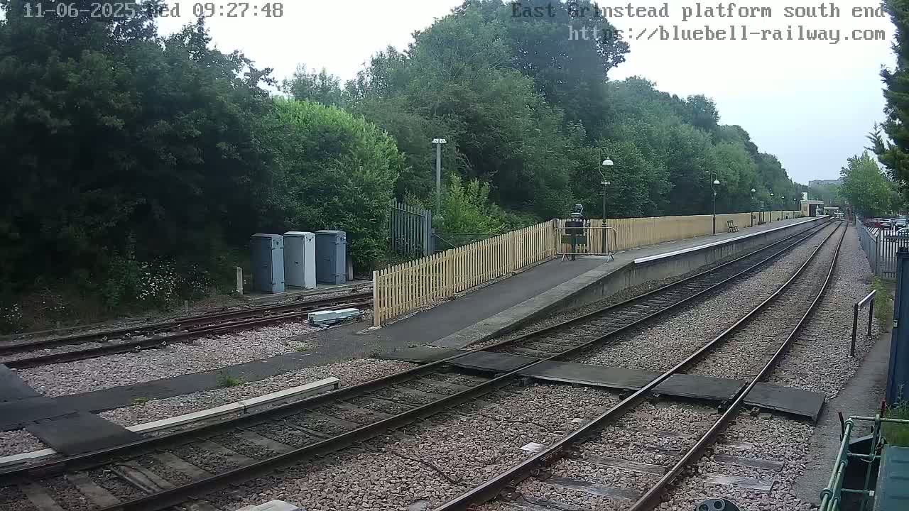 A view of a train station platform on an overcast day, showing train tracks extending into the distance, bordered by a wooden fence and lush green trees.