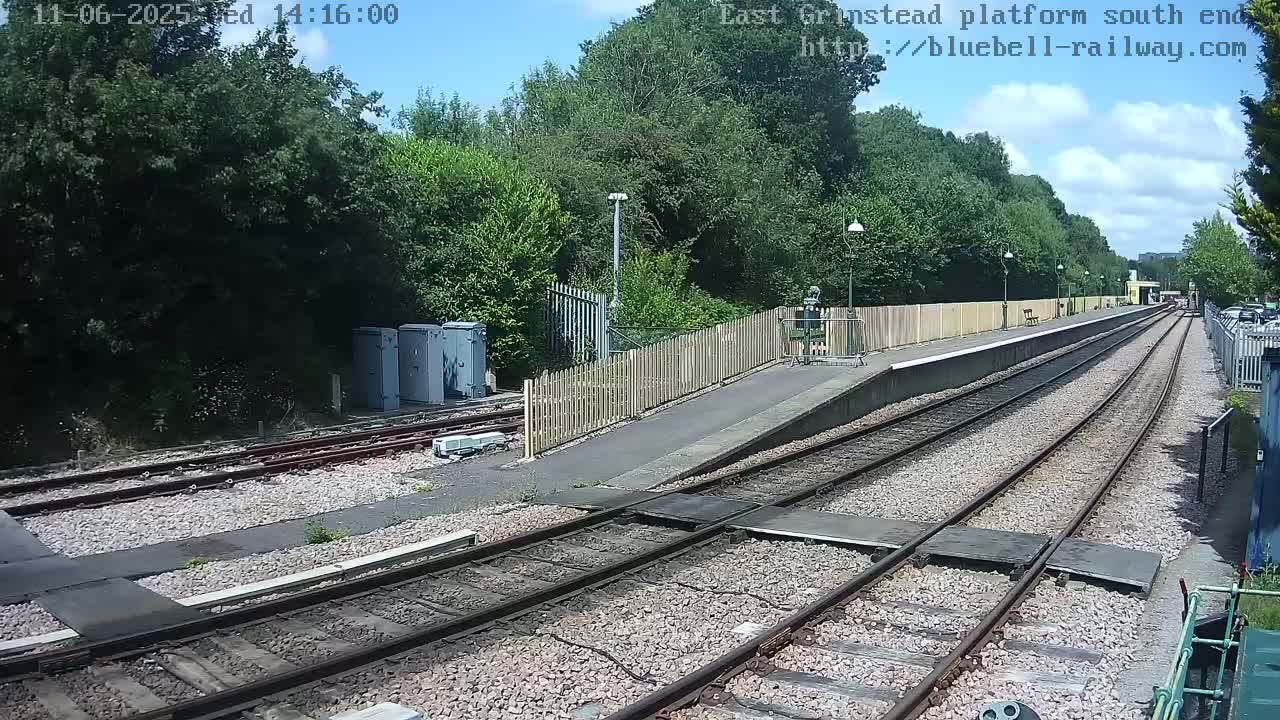 A sunny day at a train station shows a view down two train tracks that lead to a platform bordered by a wooden fence and lush green trees.