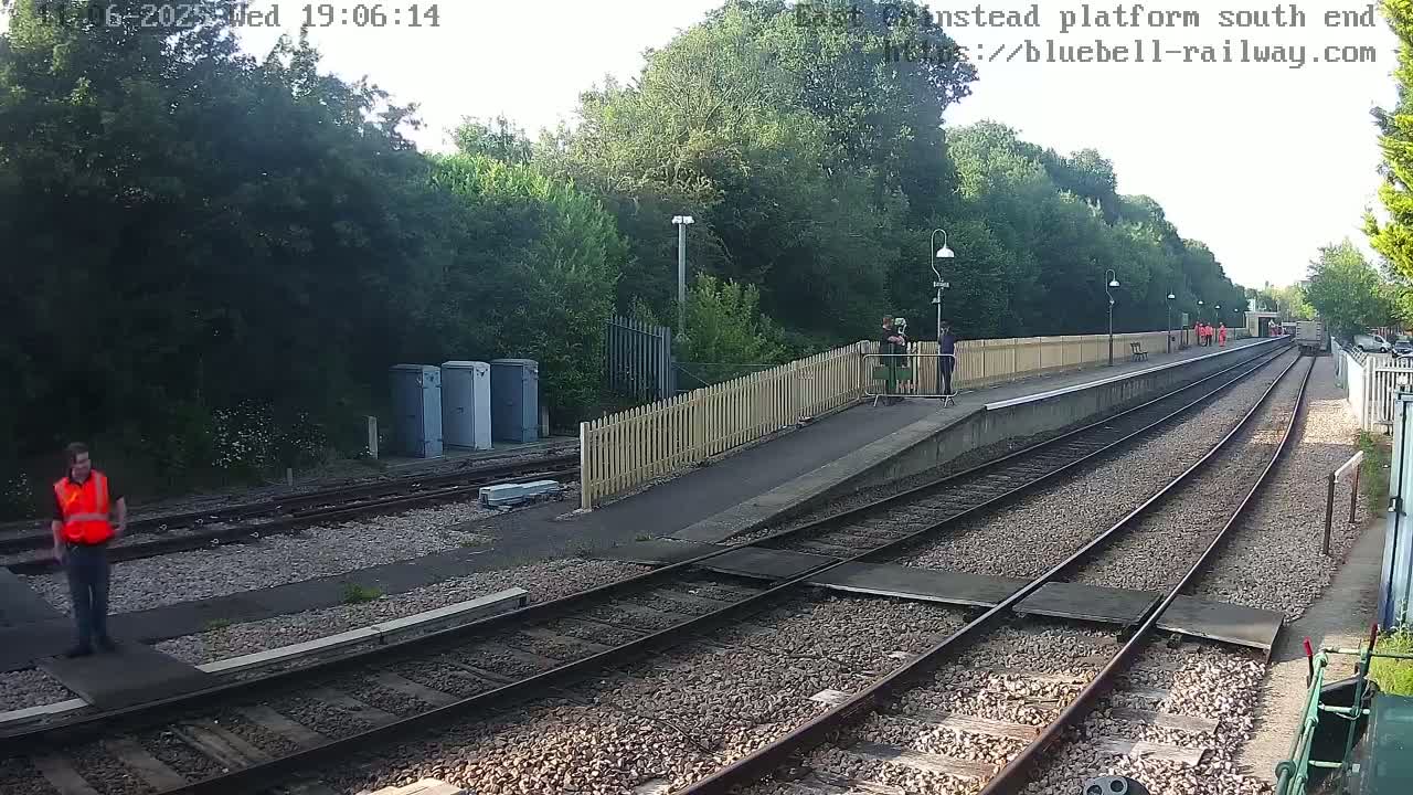 A person in a bright orange safety vest stands on a railway platform next to train tracks on a partly sunny day.