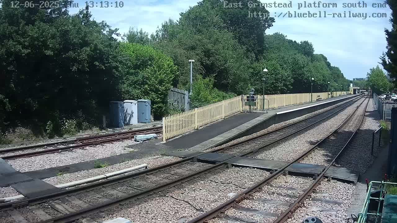 A railway platform with two sets of train tracks extends into the distance under a partly sunny sky, surrounded by lush green vegetation.