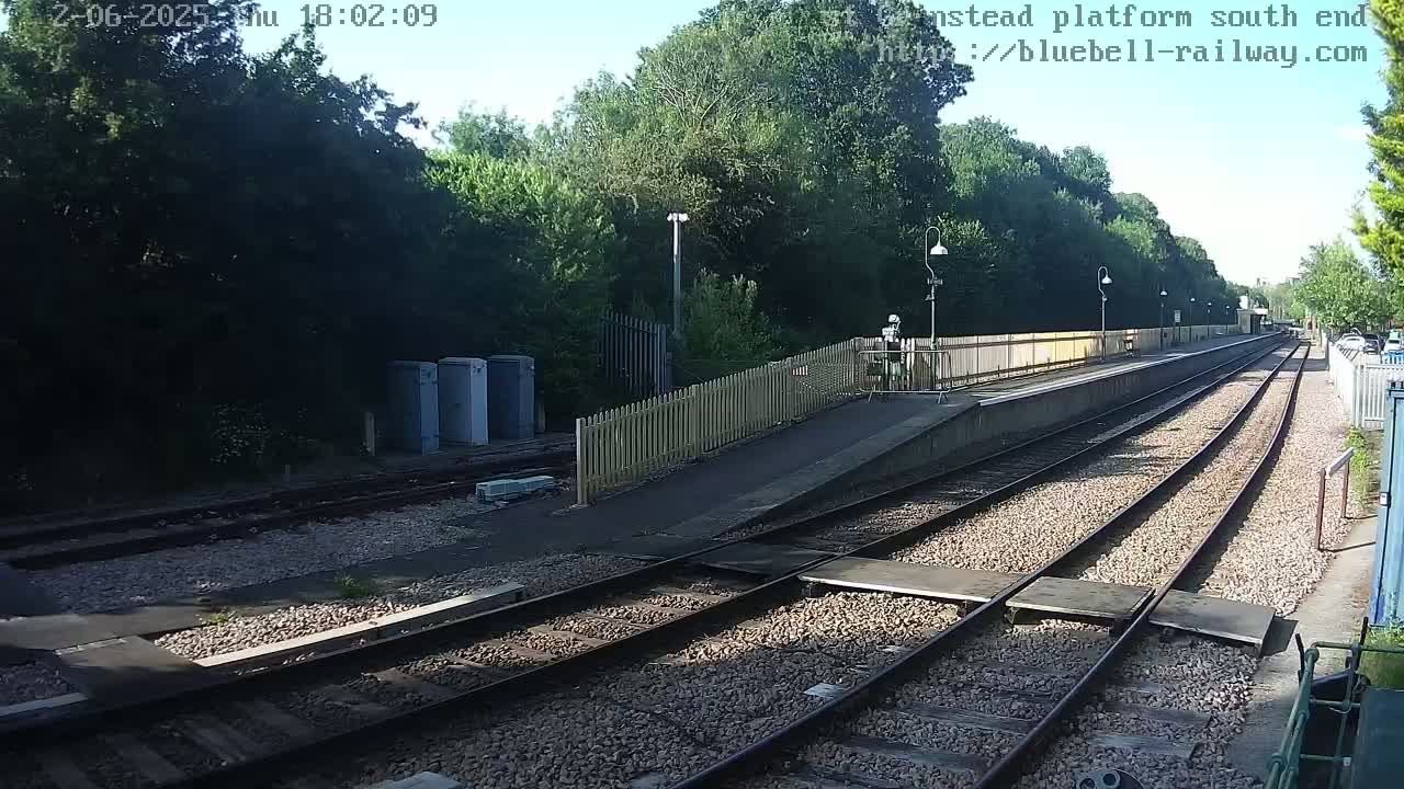 A sunny view of a train platform with two sets of tracks extending into the distance, bordered by a wooden fence and lush green trees.