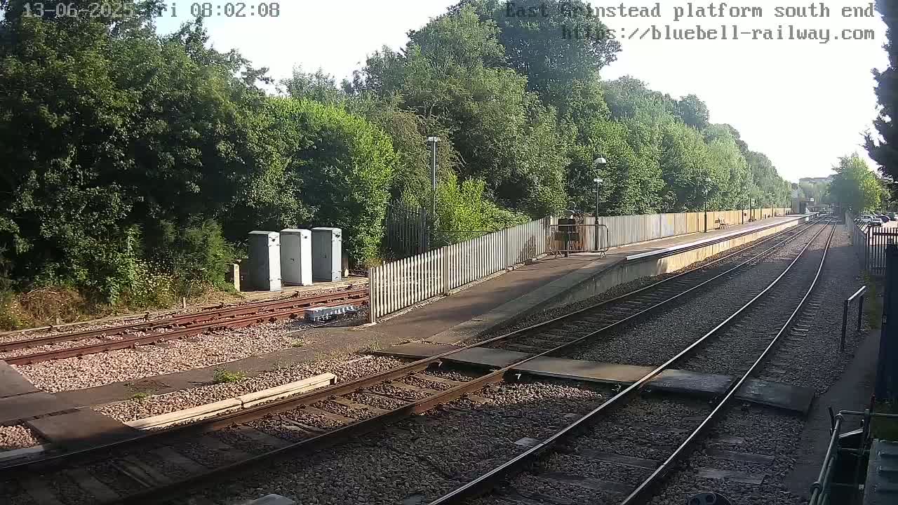 A sunny day at a train station platform with multiple train tracks, a wooden fence, and lush green trees in the background.