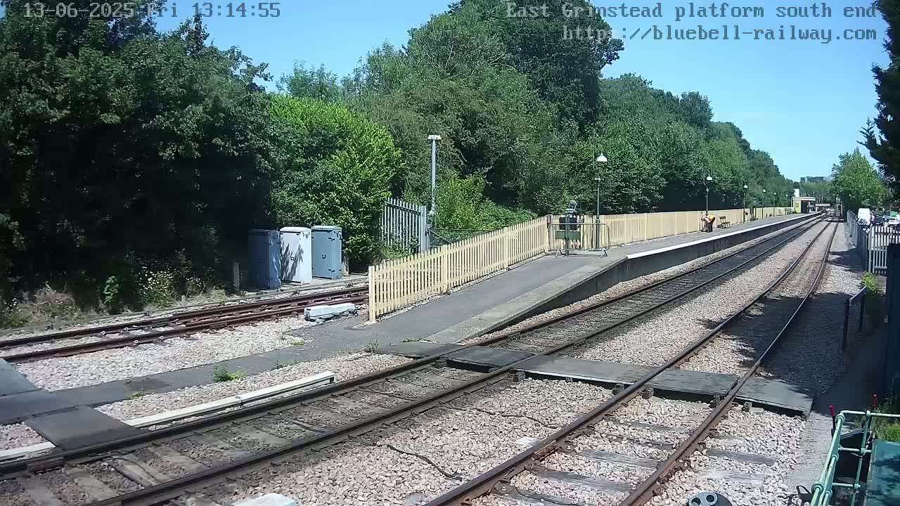 A mostly sunny view of a train platform with two sets of tracks, a wooden fence, and several utility boxes beside the tracks.