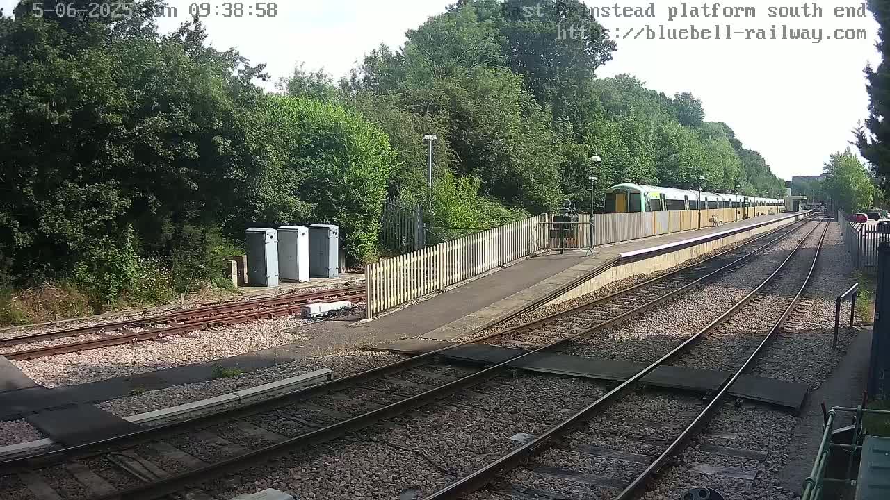 A train is stopped at a station platform next to a wooded area on a sunny day.