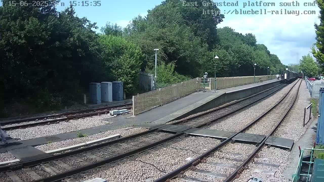 A train departs a railway platform under a partly sunny sky, surrounded by lush green trees and shrubbery.