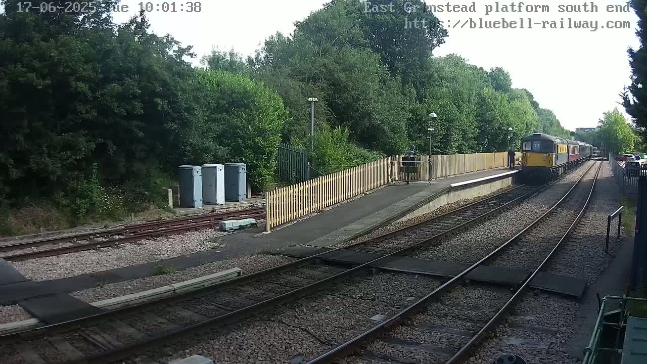 A diesel train departs a railway platform on an overcast day, passing alongside a wooden fence and lush green trees.