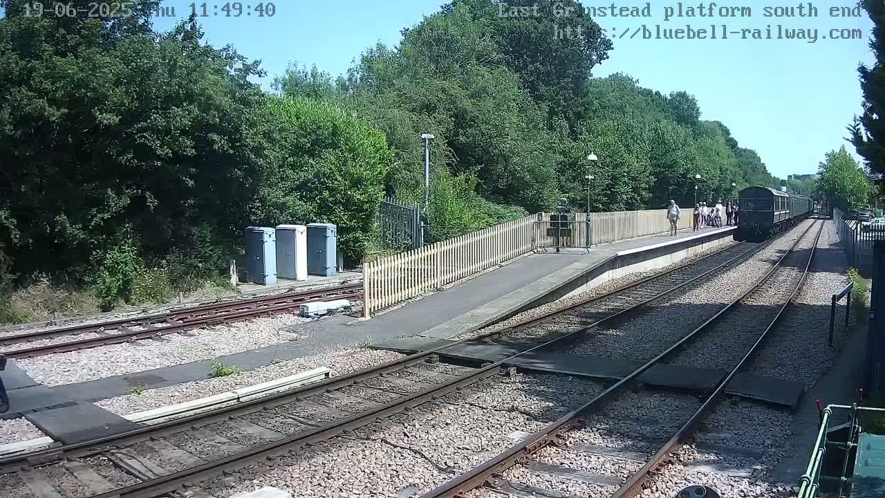 A dark-colored train is at a railway platform on a sunny day, with several people standing nearby.