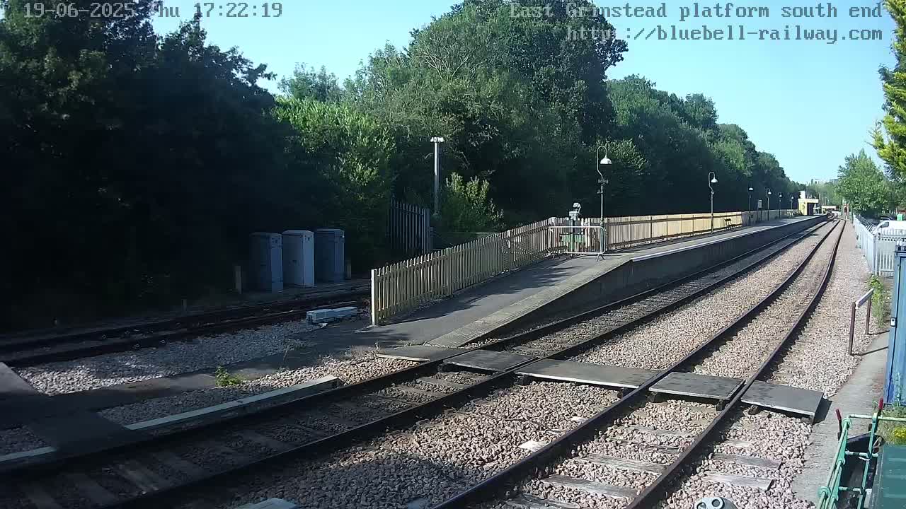A train platform with multiple train tracks and a wooden fence, under a mostly sunny sky.