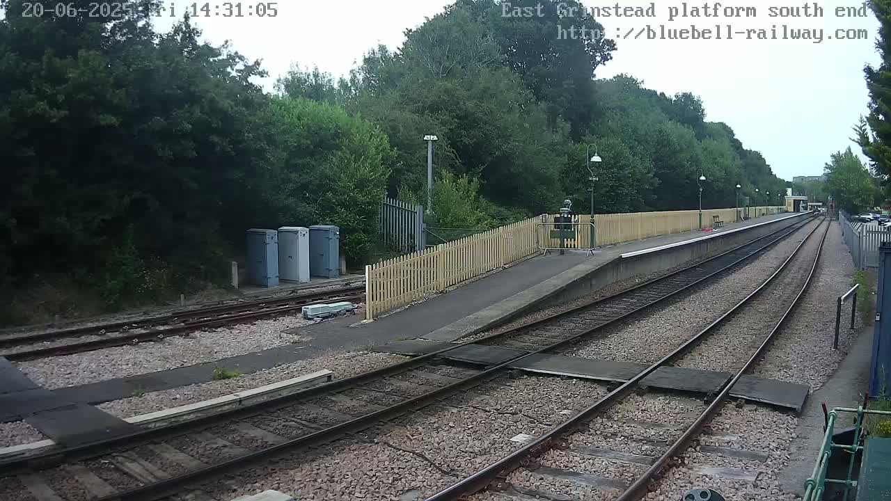 A train platform with two sets of tracks extends into a wooded area under an overcast sky.