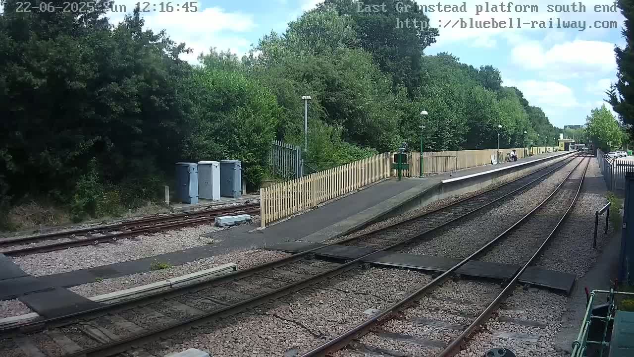 A train platform with two tracks extends into a wooded area under a partly sunny sky.
