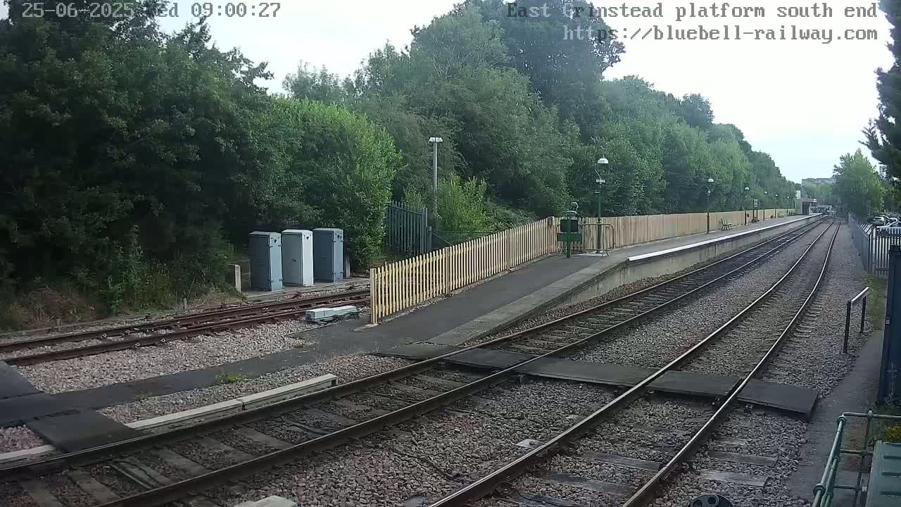 A train platform with a wooden fence, several porta-potties, and two sets of train tracks extends into a wooded area under an overcast sky.
