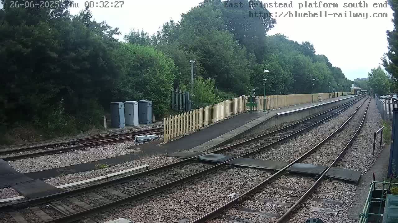 A view of a train platform on an overcast day, showing train tracks extending into the distance, bordered by a wooden fence and lush green trees.