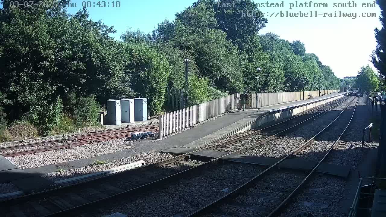 A sunny day at a train station platform with two sets of train tracks and a wooden fence bordering lush green trees.