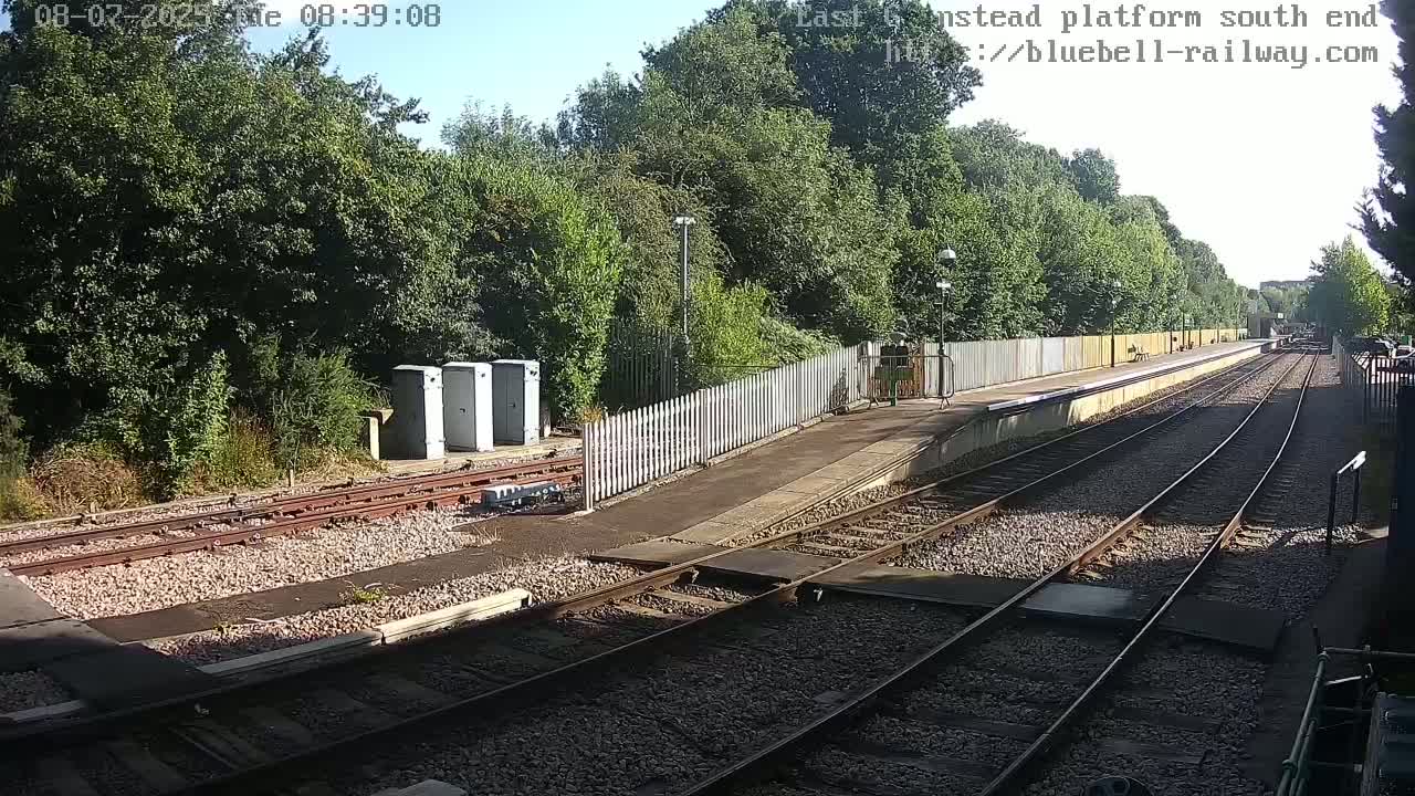 A sunny view of a train platform with multiple train tracks leading to a lush green wooded area beyond a fence.