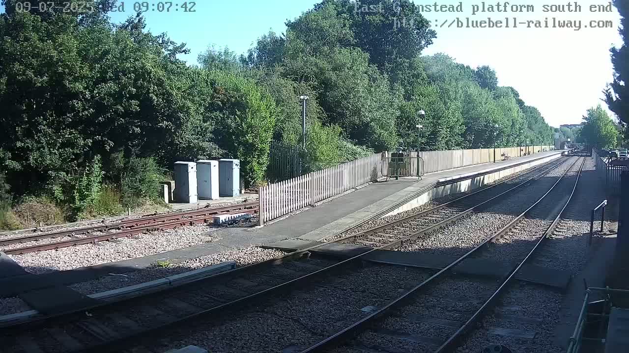 A sunny view of a train platform with multiple tracks, a wooden fence, and several small buildings beside lush green trees.