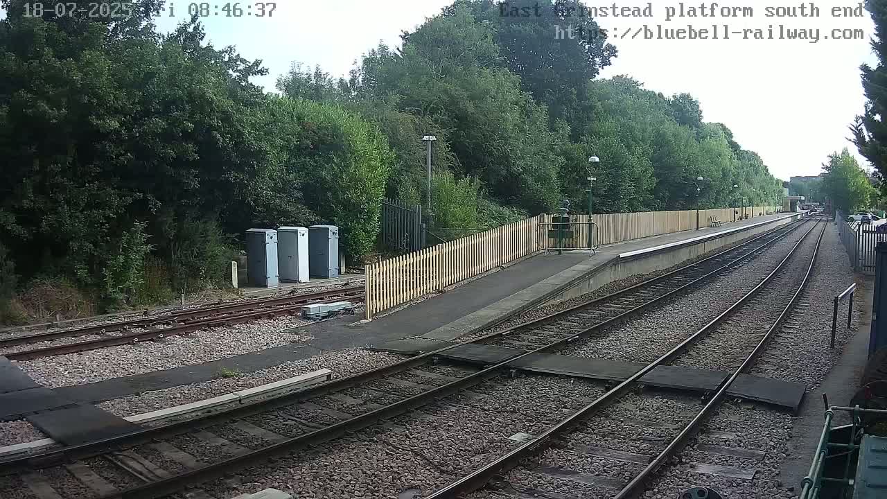 A view of a train platform on a partly cloudy day, showing train tracks extending into the distance, bordered by a wooden fence and lush green trees.