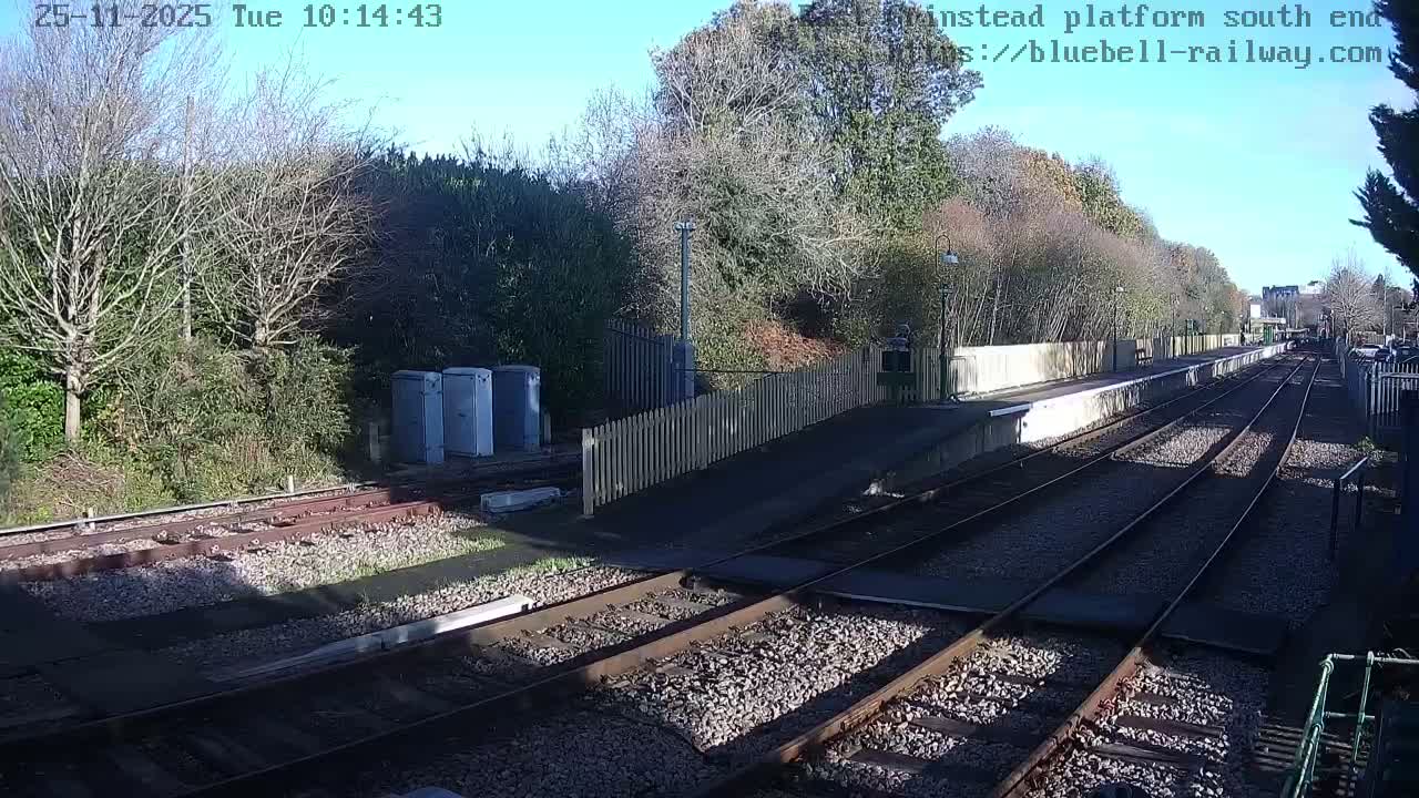 A railway scene unfolds with multiple tracks and a station platform featuring a white picket fence, all bathed in sunlight under a clear blue sky, with a mix of bare and evergreen trees lining the perimeter on a dry, late autumn or early winter day.