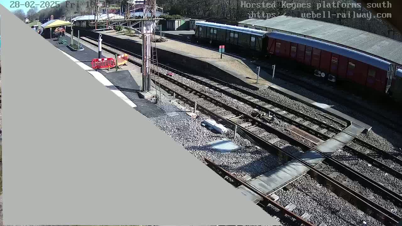 A sunny day at a train station shows railway tracks, a train car, and a platform with a small shelter.