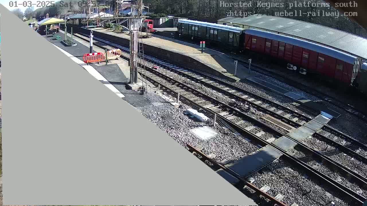A sunny day at a train station shows railway tracks, train cars, and a platform with a small building and construction barriers.