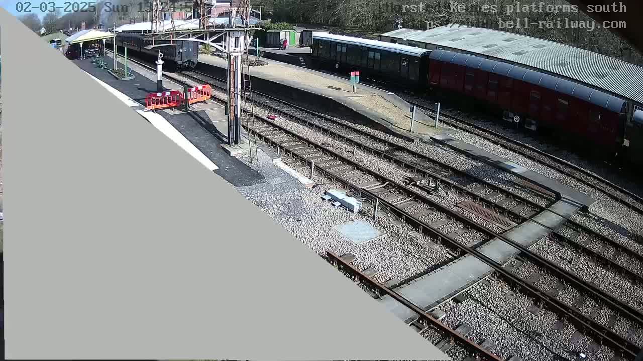 A sunny day at a train station shows several train cars parked on the tracks, with a partially visible platform and a waiting shelter.