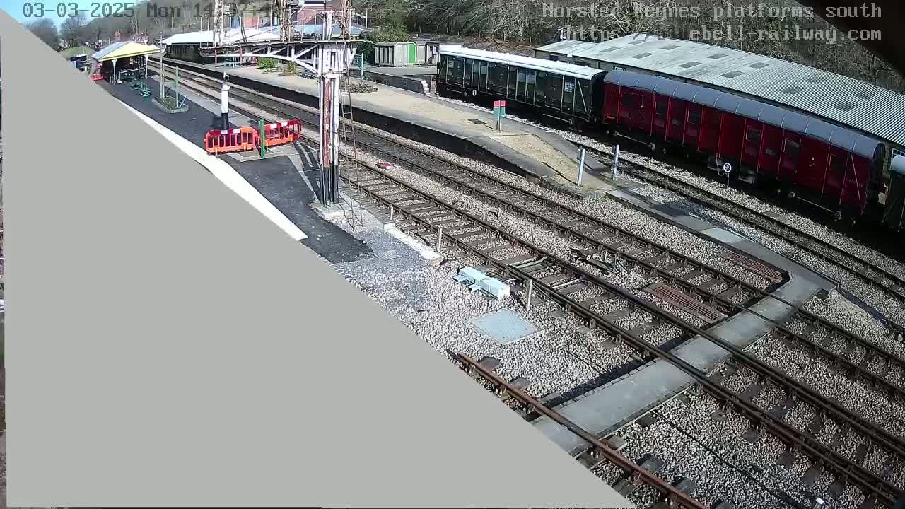 A sunny day at a train station shows railway tracks, a red train car, and a passenger train.