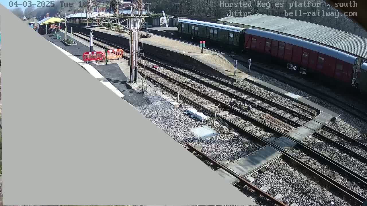A sunny day at a train station shows railway tracks intersecting, with passenger and freight cars parked nearby, and a platform with a small shelter.