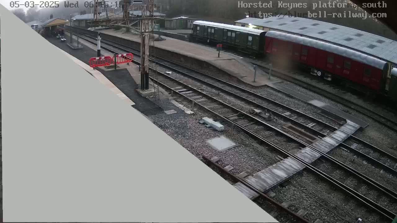 A high-angle view of a train station platform with railway tracks and two passenger rail cars, one green and one red, under overcast skies.