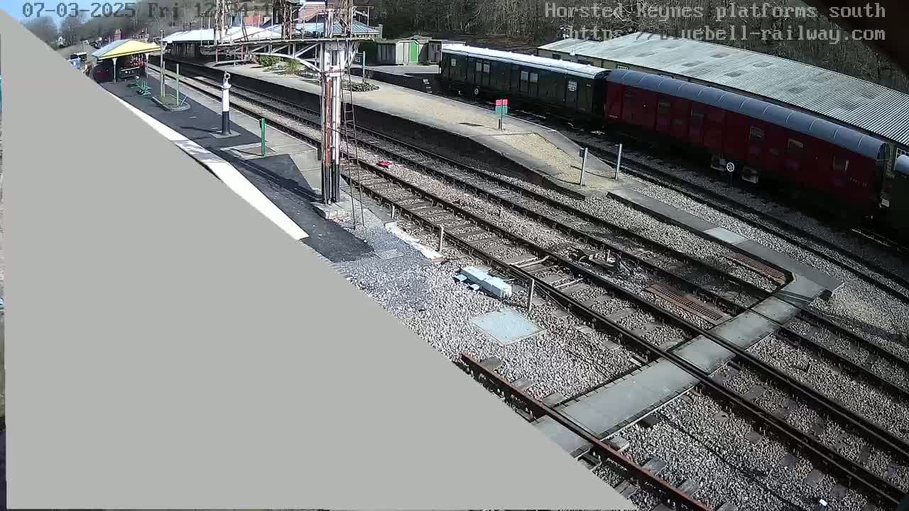 A train station on a sunny day with several train cars visible on the tracks, and a platform with a covered waiting area.