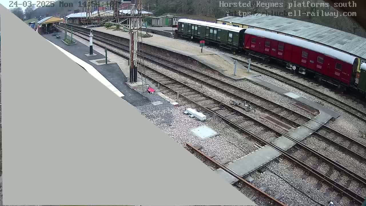 An overcast day at a train station shows several railway cars parked along multiple train tracks, with a covered waiting area visible in the background.