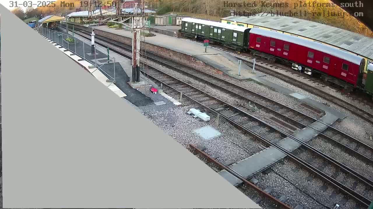 An overhead view of a train station with multiple railway tracks, a red and a green train car, and a partly constructed platform under an overcast sky.