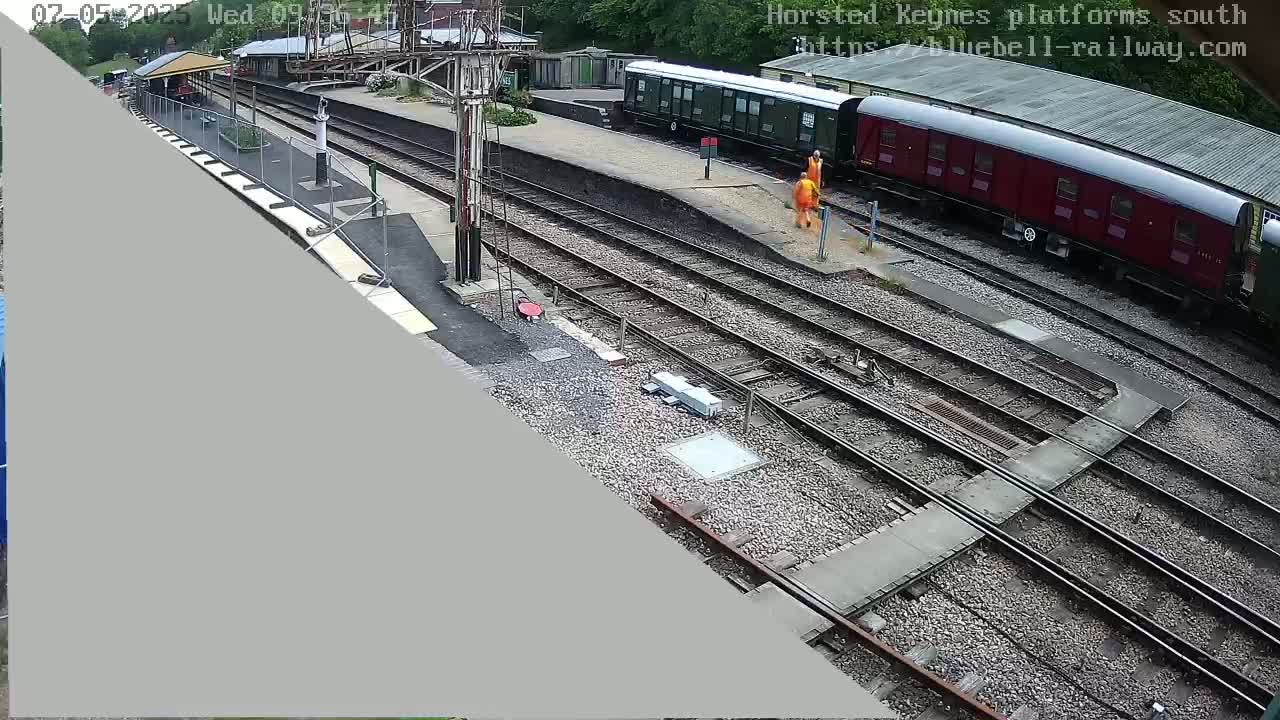 A high-angle view shows a train station with railway tracks, a partially visible train, and two people in orange vests near the tracks on a partly sunny day.