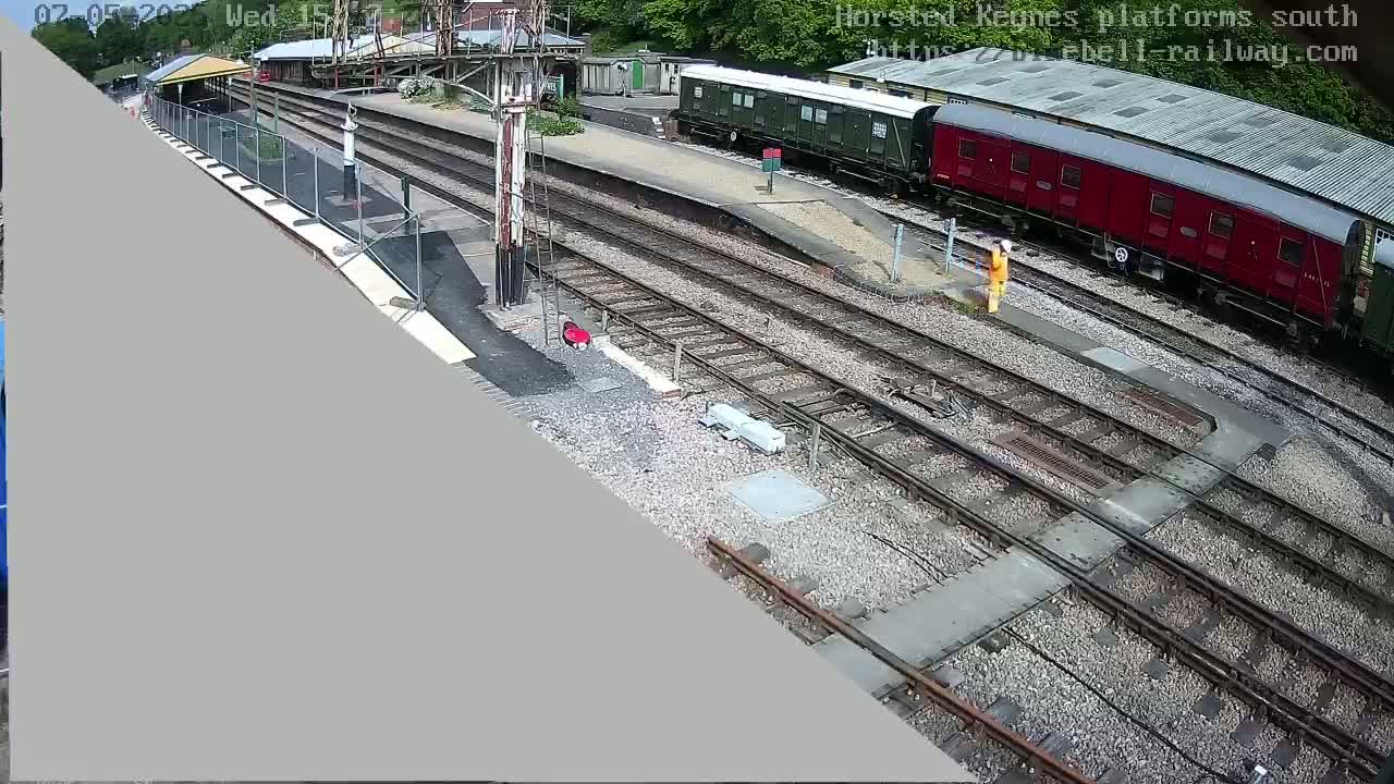 A high-angle view shows a railway station with multiple train tracks, a partially constructed platform, and two railway carriages, one dark green and one dark red, under a partly sunny sky.