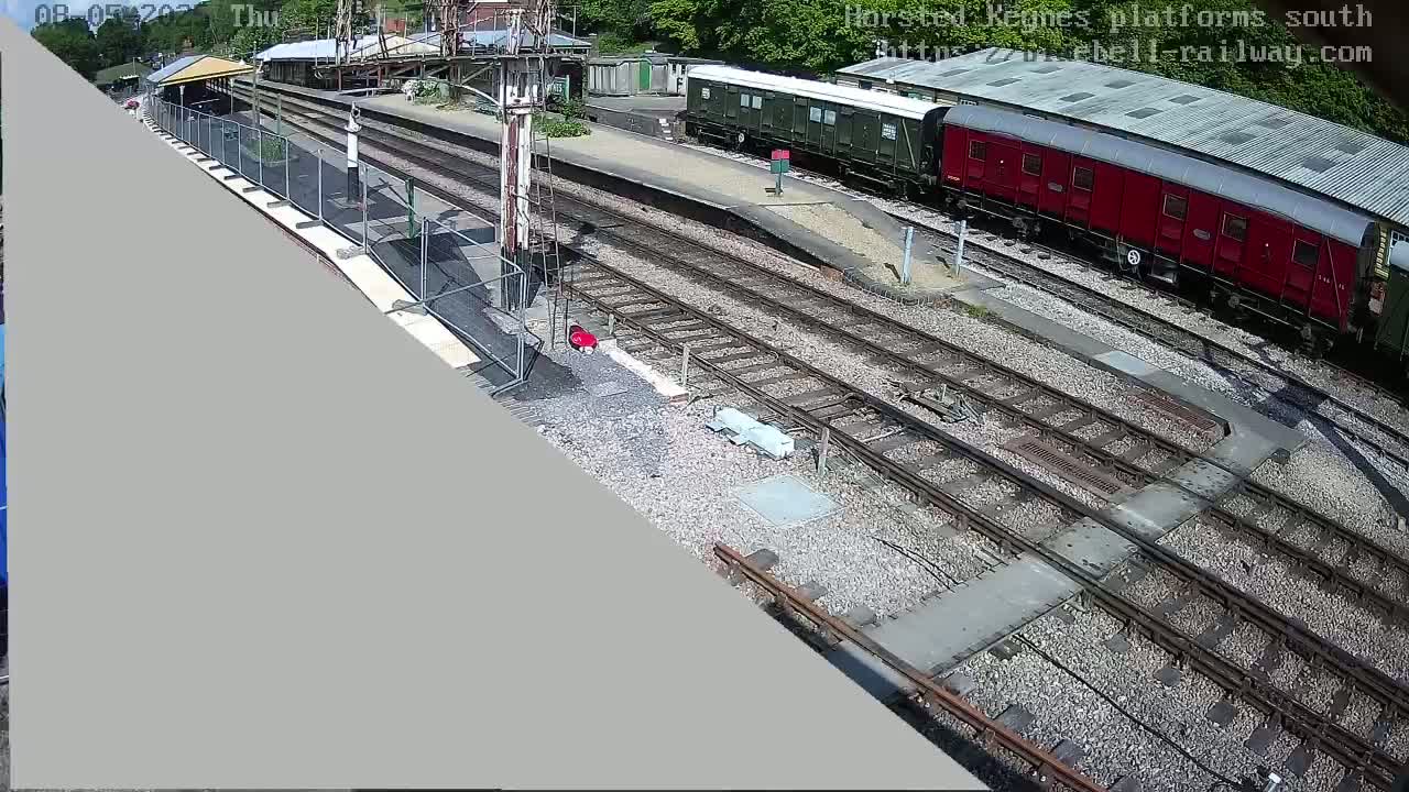 A high-angle view shows a railway station on a sunny day, with several train cars parked on the tracks and a person in red near a platform.