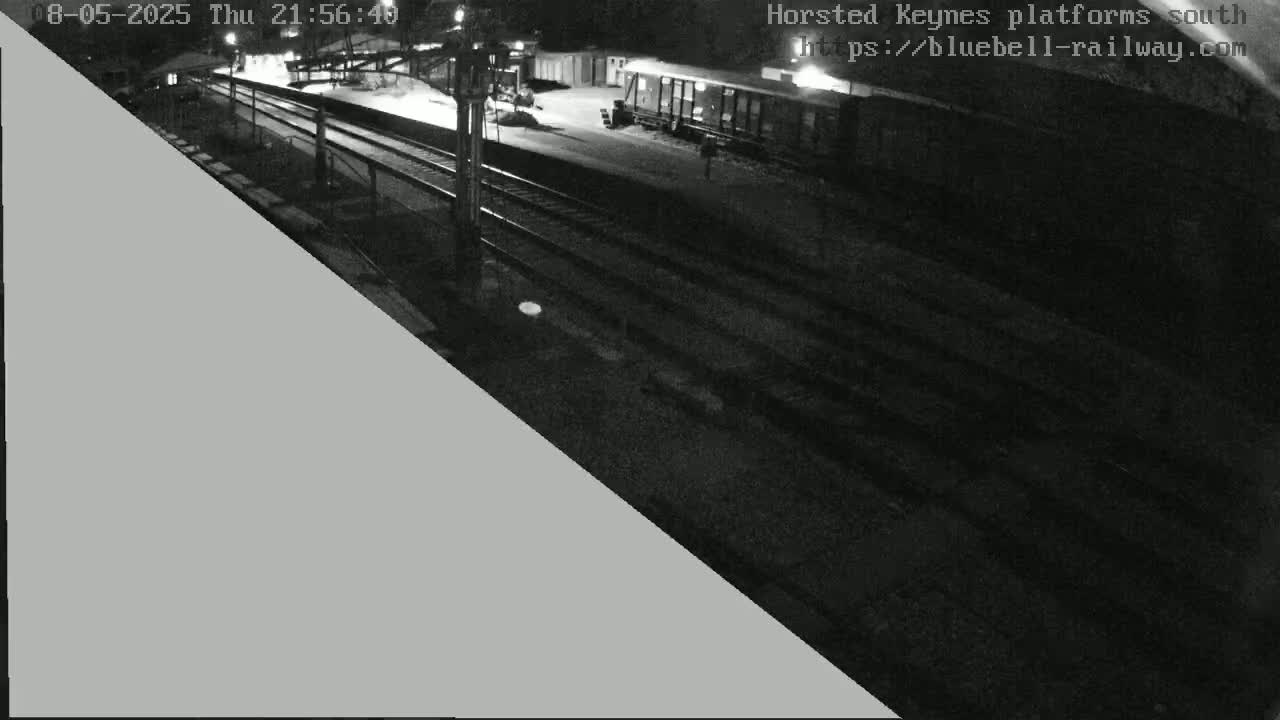 A nighttime view of a train station platform with railway tracks and a train car visible under artificial lighting.