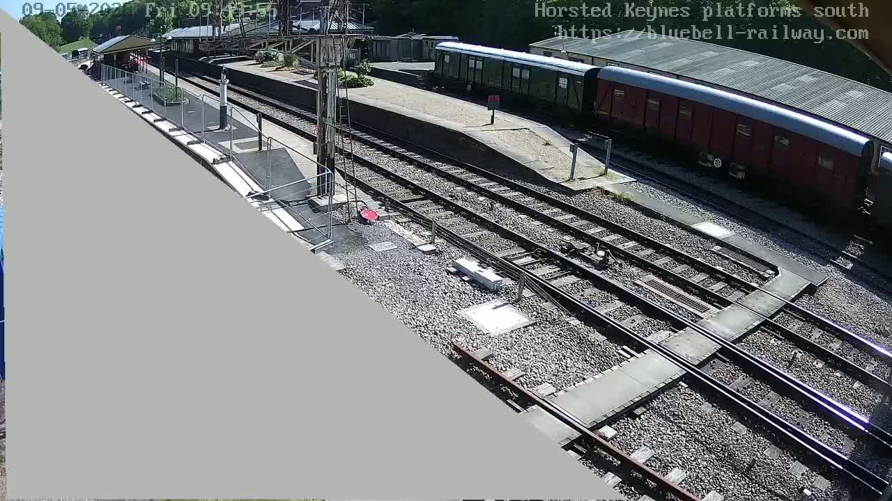 A sunny overhead view of a train station platform with railway tracks, train cars, and a building.