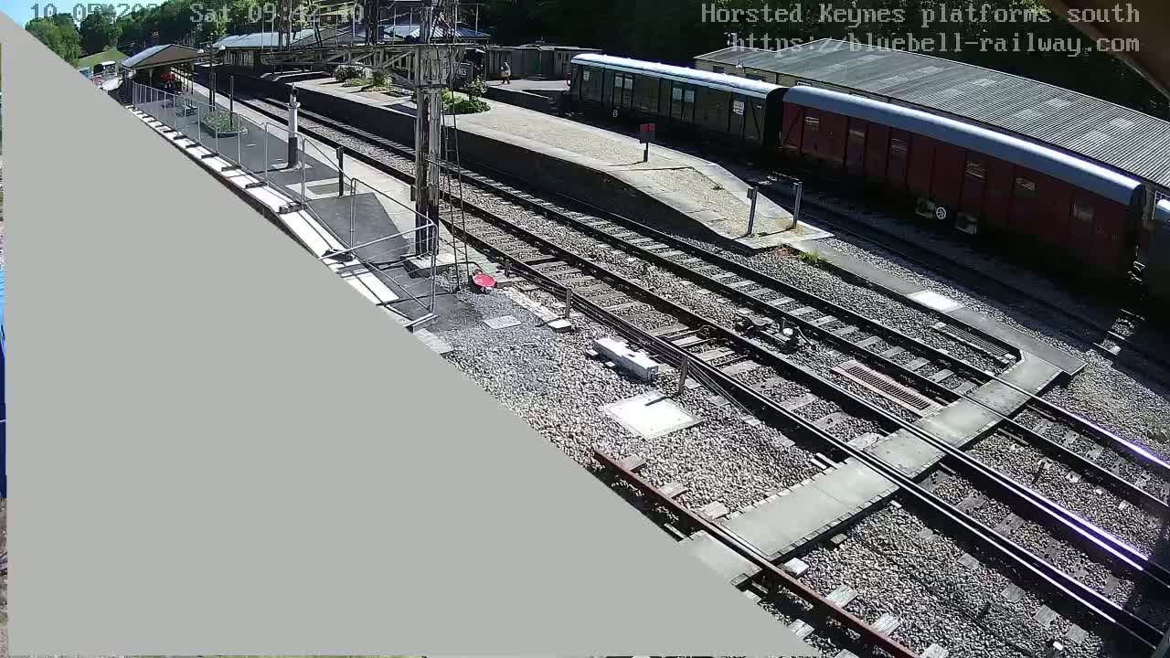 A high-angle view of a railway station platform on a sunny day, showing multiple train tracks and railway cars.