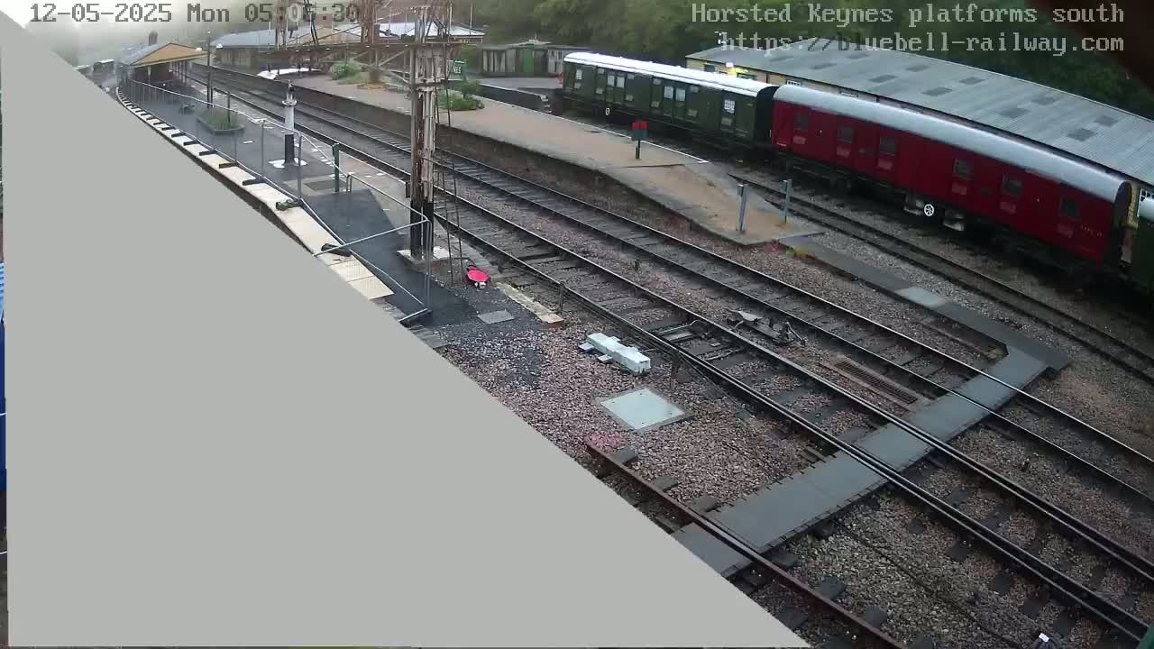 A high-angle view shows a railway station with multiple train tracks, a red and a green train, and a small station building under an overcast sky.