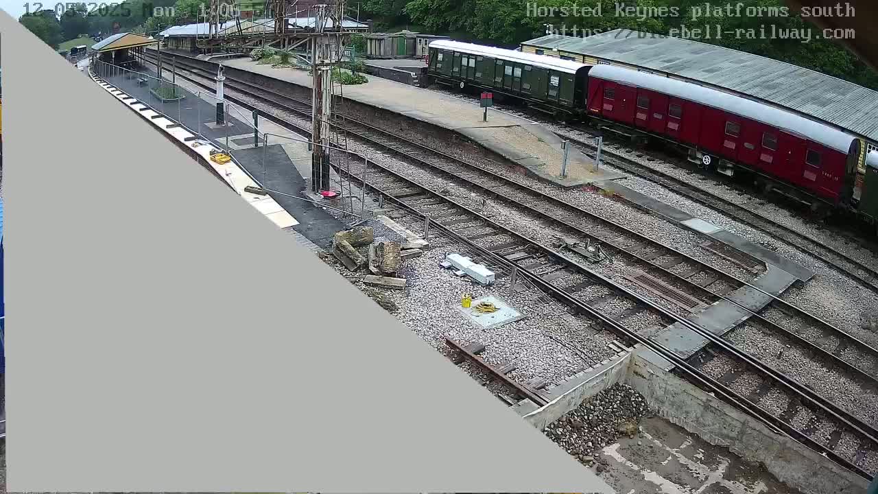 A high-angle view shows a train station with multiple railway tracks and two train cars, one dark green and one dark red, parked on the tracks in an outdoor setting on an overcast day.