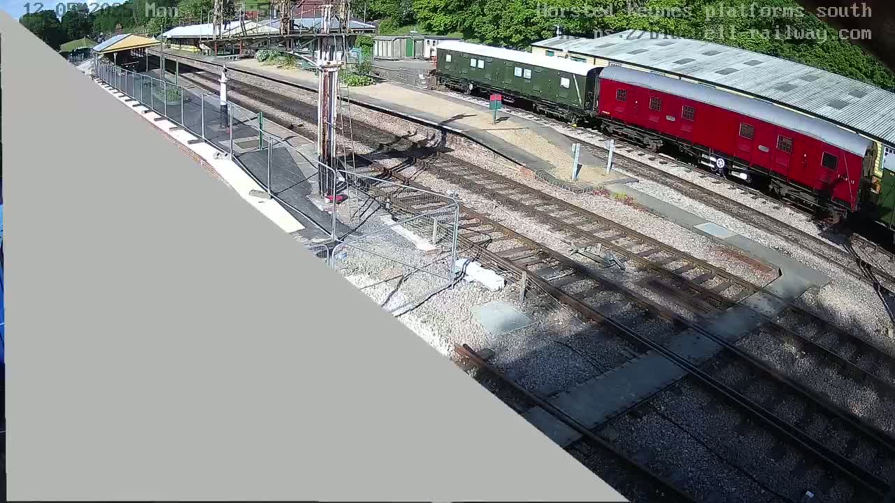 A high-angle view shows a railway station with a green and a red train car on the tracks under a sunny sky.