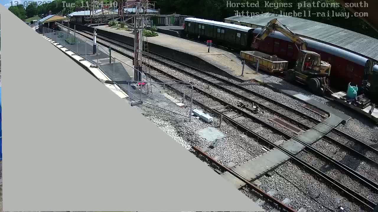 A railway platform under construction on a sunny day, with a train, excavator, and construction materials present.