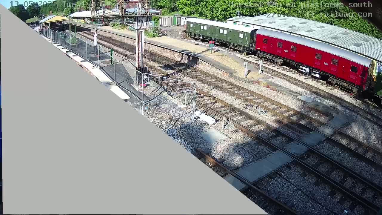A high-angle view of a railway station on a sunny day shows several train cars parked on the tracks near a platform with construction barriers.