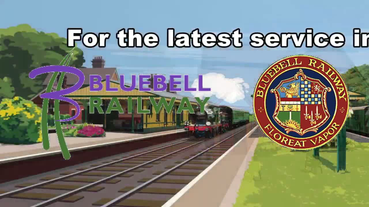 A steam train pulls into a station on a sunny day, passing lush green landscaping.