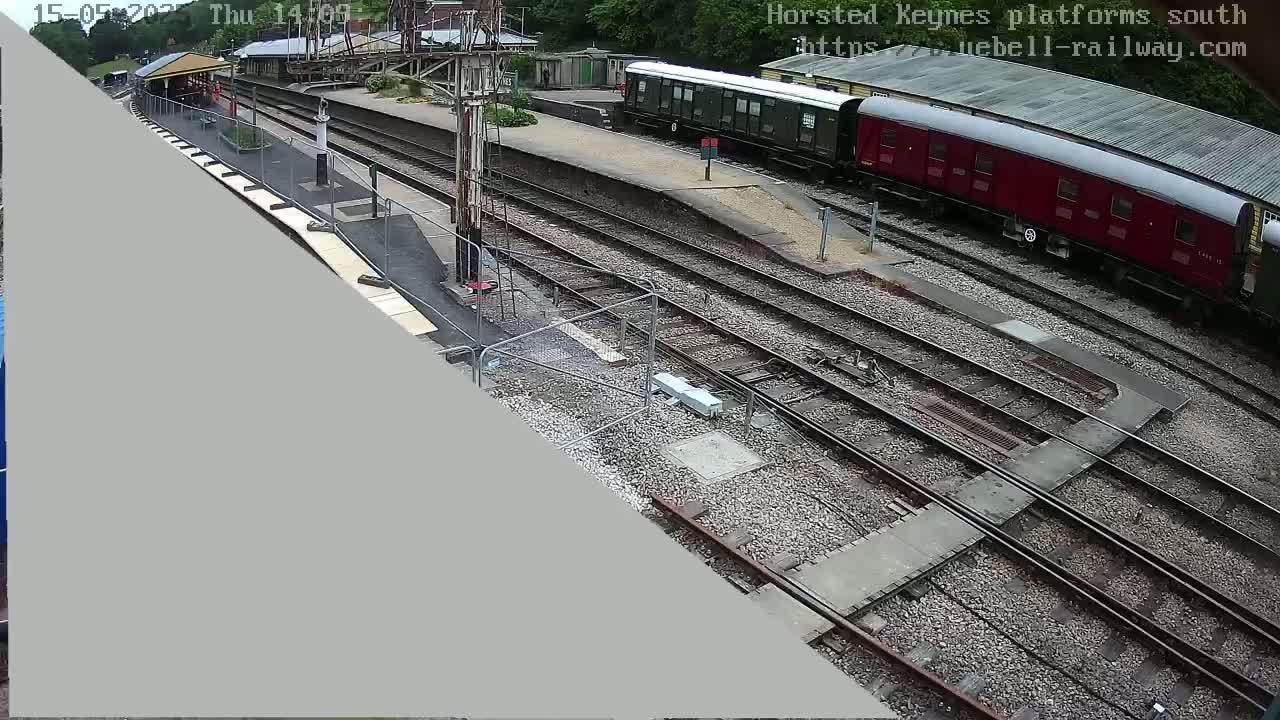 A high-angle view shows a railway station with multiple train tracks, a partially visible train, and a small section of a platform, under a partly cloudy sky.