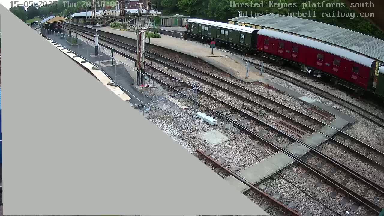A high-angle view shows a railway station with multiple train tracks, a partially-constructed platform, and two passenger cars parked on the tracks.