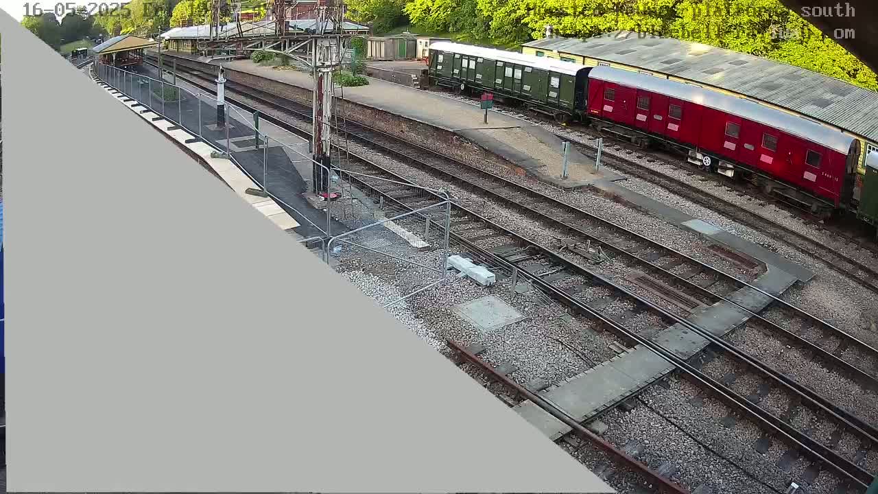 An overhead view of a train station on a sunny day shows several railway cars, including a long red one, parked on multiple tracks.
