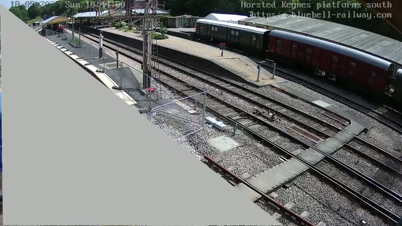 A high-angle view of a train station on a sunny day shows multiple train tracks, a partially visible train, and a platform with a fence.