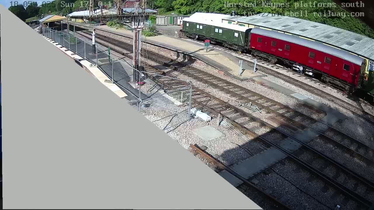 A high-angle view shows a railway station on a sunny day with a red and a green train car parked on the tracks.